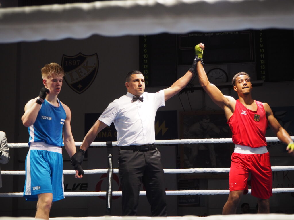 Tuomari nostaa punaiseen asuun pukeutuneen nyrkkeilijän käden ylös voiton merkiksi.