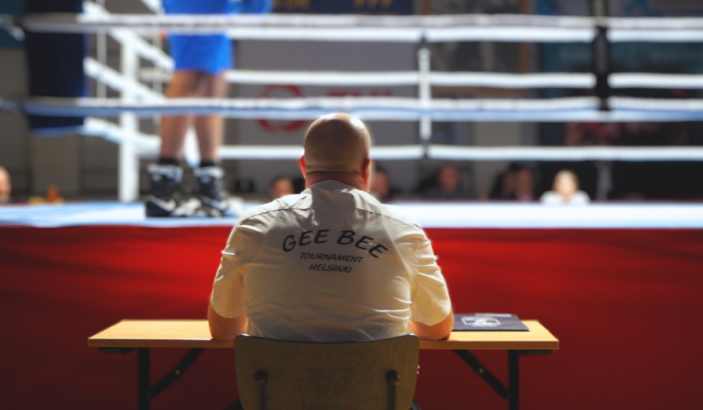Nyrkkeilytuomari istuu selin kameraan, taustalla nyrkkeilykehä. Tuomarin paidan selässä teksti GeeBee Tournament Helsinki.