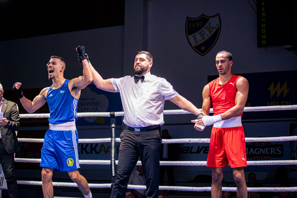 Kaksi nyrkkeilijää kehässä. Tuomari nostaa sinisessä asussa ottelevan nyrkkeilijän käden ylös voiton merkiksi.