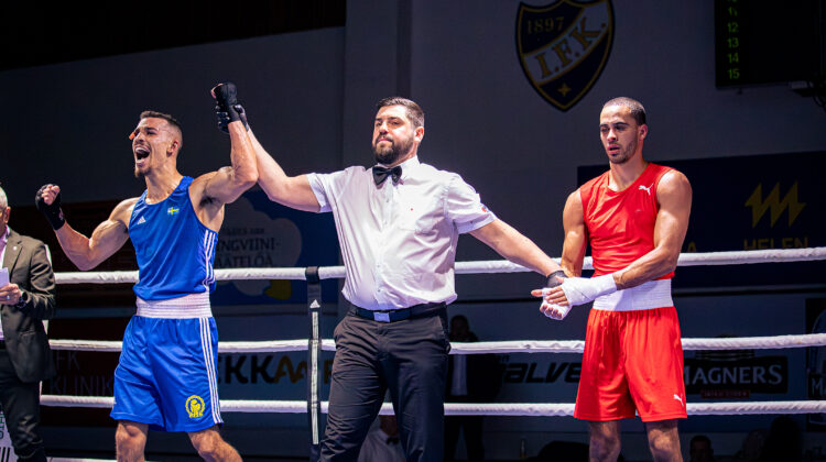 Kaksi nyrkkeilijää kehässä. Tuomari nostaa sinisessä asussa ottelevan nyrkkeilijän käden ylös voiton merkiksi.