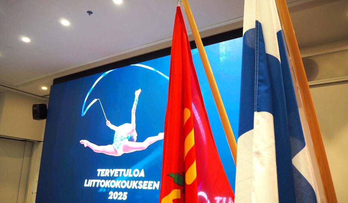 Taustalla dia, jossa voimistelija hyppää nauhan kanssa sekä teksti Tervetuloa liittokokoukseen 2025. Etualalla punainen TUL:n lippu sekä Suomen lippu.