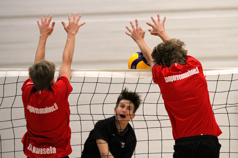 Lentopallon pelitilanne. Kaksi poikaa punaisissa paidoissa on torjumassa palloa, kun toisella puolelta on tulossa iskulyönti.