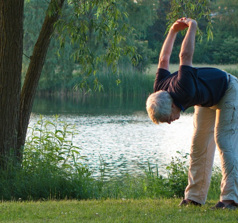 Seniori-ikäinen mies venyttelee järven rannalla nurmikolla.