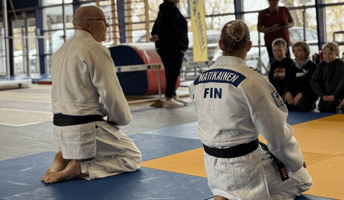 Kaksi judokaa istuu selin lapsiryhmän edessä judomaton päällä. Taustalla näkyy lapsia istumassa sekä aikuisia seisomassa.