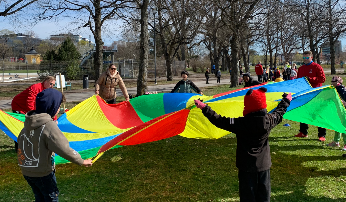 Lapsia ja aikuisia leikkimässä värikkäällä leikkivarjolla ulkona.