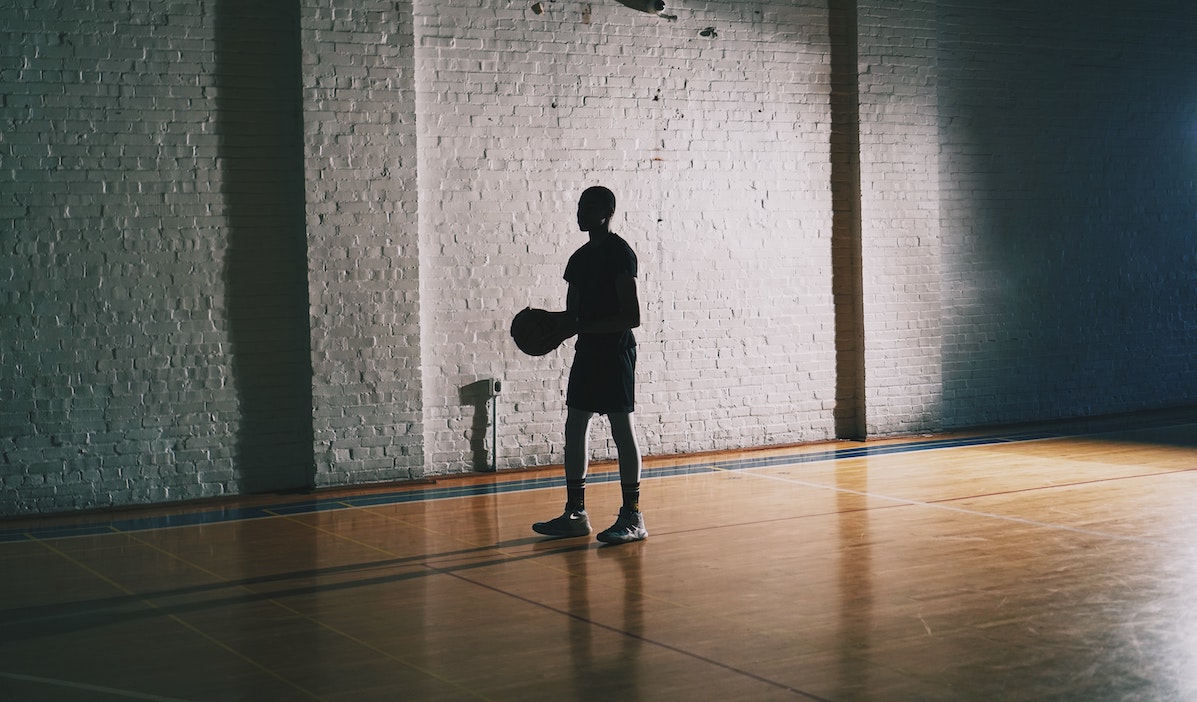 Pimeässä liikuntasalissa yksinäinen poika pitelee koriapalloa käsissään.