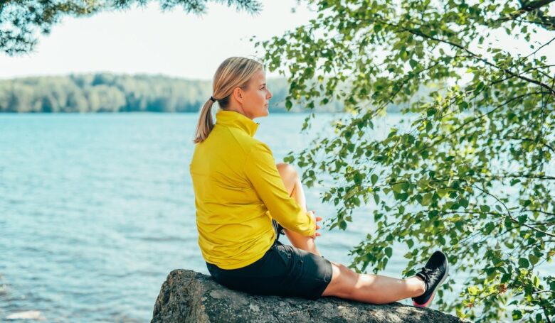 Nainen istuu kivellä keltaisessa paidassa katsellen järvelle