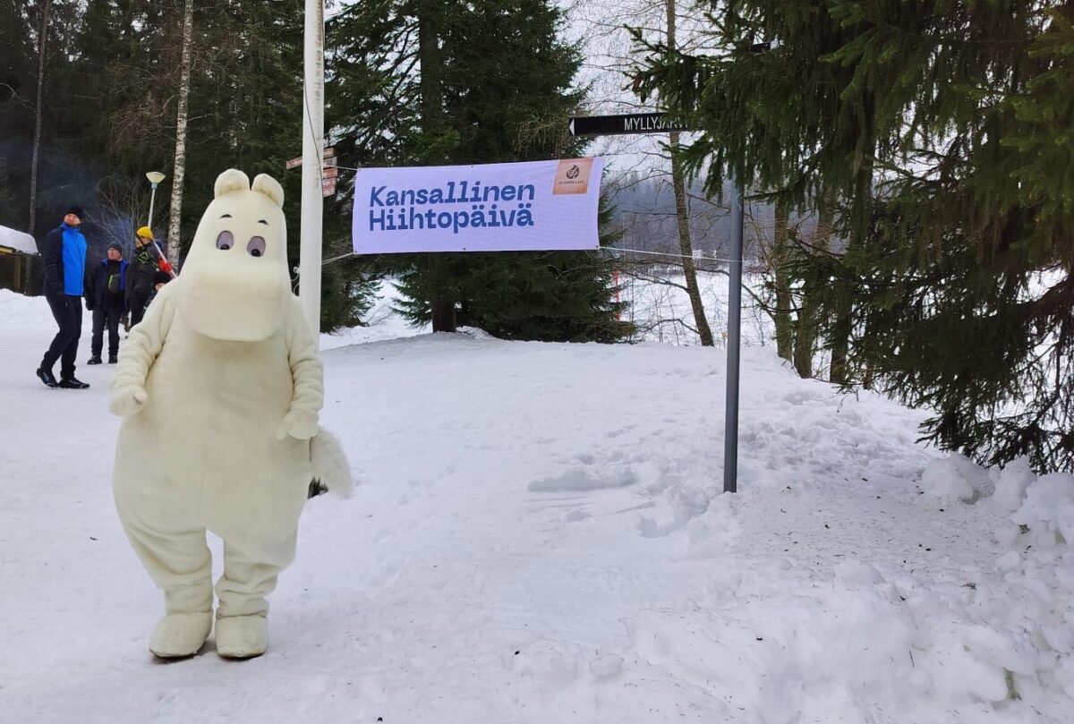 Muumipeikko seisoo lumisessa maisemassa, taustalla banderolli jossa teksti Kansallinen Hiihtopäivä.