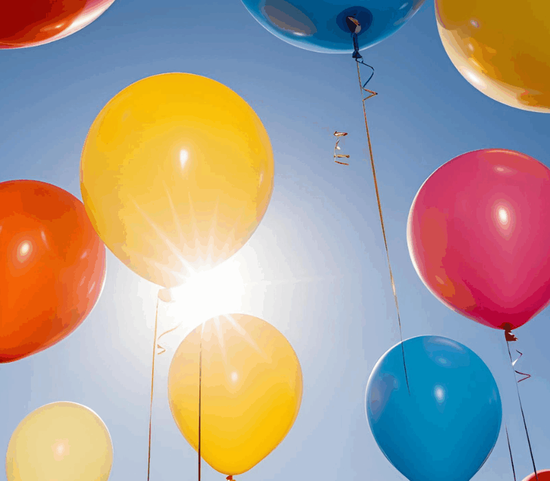 Monivärisiä ilmapalloja taustallaan sininen taivas. Keskellä kuvaa näkyy myös aurinko.