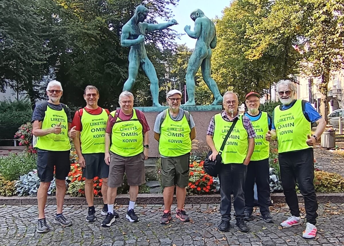 Kävelypässit eli seitsemän seniori-ikäistä miestä huomioliivit päällään seisomassa nyrkkeilijäpatsaan edessä ulkona.