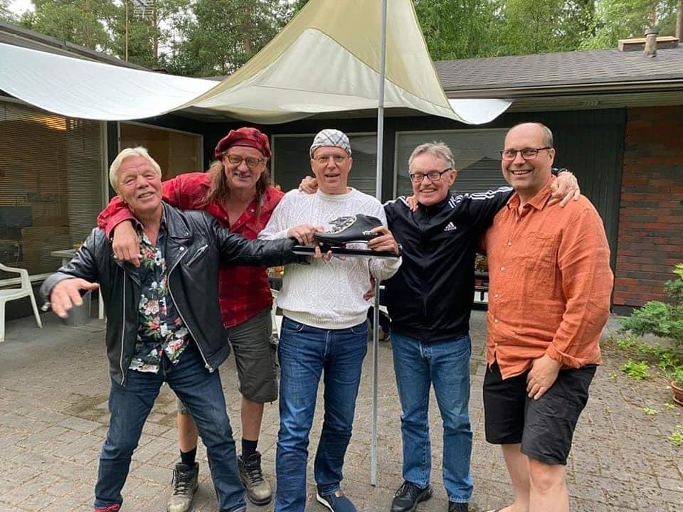 Viisi miestä seisoo rinnatusten iloiset ilmeet kasvoillaan. Keskimmäisen kädessä pikaluistin.