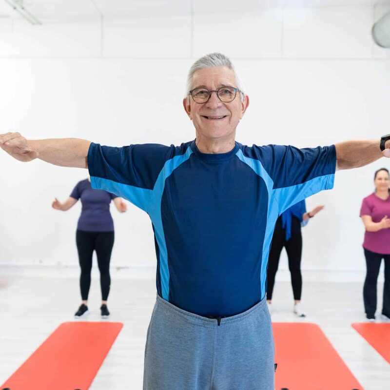 Seniori-ikäinen mies jumppaa kädet levitettynä, taustalla jumppamatoilla kolme henkilöä.